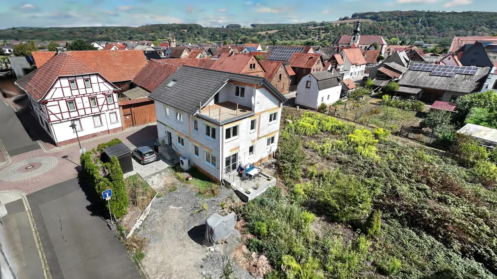 Ein Zweifamilienhaus mit Garten, dahinter Nachbarschaft und Hügel, in der begehbaren 3D-Außenansicht aus Drohnenaufnahmen